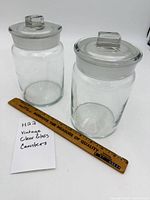 Both clear glass canisters with lids beside ruler for scale