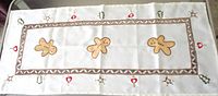 Full view of white silk table runner with three gingerbread men, geometric border, embroidered motifs