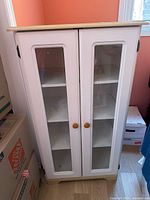Front view of white cabinet showing glass doors, knobs, shelves, wood top and base.