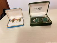 Both presentation cases open showing rectangular jade cufflinks in green Dante box and oval cufflinks with tie clip in blue case