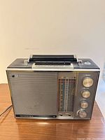 Front view showing speaker grille, tuning scale, control knobs, handle, antenna and attached AC cord