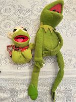 Both Kermit plush toys side by side on lace surface