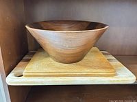 Walnut-tone wooden salad bowl resting on stacked light-colored wooden cutting boards inside shelving