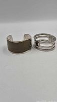 Two silver bracelets shown side by side: wide inlaid cuff and open-bar hinged design