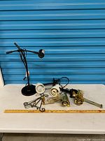 Group shot of entire lot on table: desk lamp, two white clamp lights, brass hardware, metal stands, ruler for scale