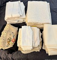 Stacked piles of cream, white and black table linens including lace, crewel topper and napkins
