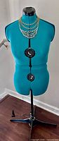 Full front view of teal Dritz SewYou dress form on black metal stand wearing gold necklace