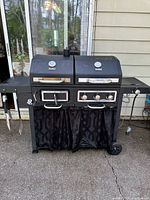 Front view of RevoAce dual fuel grill showing two cooking chambers, side burner, side shelf, utensil hooks, and base curtain