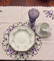 Place setting on embroidered topper showing dinner plate, salad plate, bowl, cup, saucer, purple glass