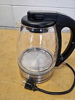 Side view of Hamilton Beach glass electric kettle showing glass body, black handle, stainless base, power cord and plug