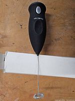 Full view of Mr. Coffee handheld frother showing handle, power button, whisk, resting on box