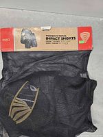 Retail package showing cardboard header and mesh bag containing black impact shorts with visible gold padded sections