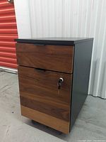 Three-quarter view showing wood veneer drawer fronts, black body, casters