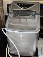 Front and side view of silver countertop ice maker with attached drain hose