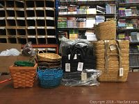 Photo showing various baskets including natural woven baskets, synthetic black baskets wrapped in plastic, and colorful small baskets in green and blue placed on a table in front of shelves.
