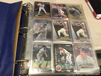 Blue 3-ring binder opened to show nine baseball cards in clear plastic sleeves, featuring various players in action.