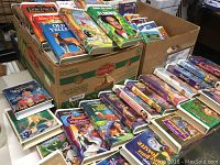Photo showing multiple VHS tapes with Disney movie covers in boxes and spread out on table.