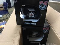 Two black boxes containing NHL Winnipeg Jets piggy banks in the shape of hockey helmets with team logo visible through the window.