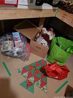 Group view showing plastic bag of gift boxes, cardboard box of fabric items, green tote bag of stockings, quilted tree wall hanging, red tote