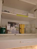 Cabinet shelf showing boxed egg cooker set, yellow cookers, decanter, flared vase