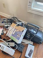 Group shot of entire electronics lot on floor: JVC boombox, two Panasonic handsets on chargers, RadioShack recorder box, power strips, cables