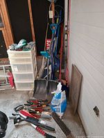 Overview of lot showing long-handled tools, snow shovels, broom head, bag of ice melt, hand tools on floor
