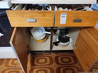 Open cabinet showing plastic colander, mixing bowls, stainless pot, small pan and open drawers with flatware