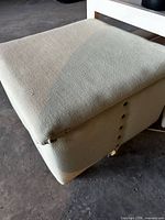 Top and side of beige linen-blend ottoman showing nailhead trim, caster foot, stain and fabric tear