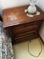angled view of brown wood nightstand showing three drawers, top surface, minor scratches, and power cord nearby
