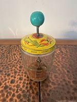 Overall view of glass butter churn showing jar, printed yellow lid, plunger mechanism and teal knob