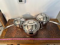 Three Belgique stainless pots with lids on wooden surface