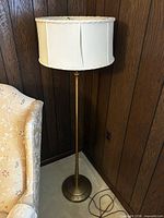 Full view of brass floor lamp next to chair showing base, pole, shade and cord
