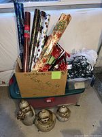 Box with wrapping paper rolls, bows, lights, plastic tote beneath, three ceramic snowmen in front