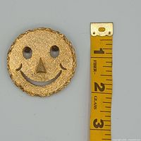 front view of gold-tone smiley face brooch beside tape for size