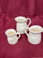 Three matching off-white ceramic creamers on burgundy fabric