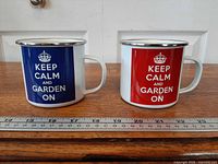Front view of both enamel mugs showing blue and red graphics and silver rims with ruler for scale