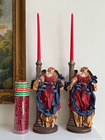 Pair of angel candle holders with red taper candles beside red bead garland in tube