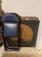 Two padded folding chairs stacked against folded card tables including ship motif table