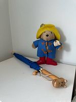 Front view of Paddington Bear plush standing beside folded blue umbrella