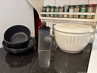 Three black bowls, stainless steel cheese grater, clear salad spinner on countertop