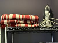 Stack of four red and white striped towels beside an electric steam iron on wire shelf