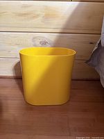 Front view of yellow rectangular plastic wastebasket on wood floor