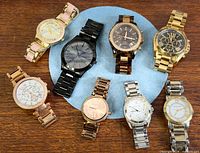Eight Michael Kors watches arranged in circle on blue pad, showing variety of styles and finishes