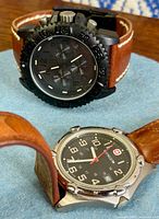 Both watches displayed on blue pad, showing faces and leather straps