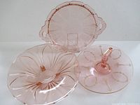 Group view of three pink depression glass pieces: handled cake plate, tidbit tray with handle, footed swirl bowl