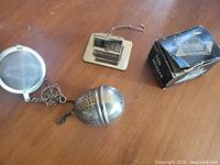 Three tea infusers laid out: mesh ball, house-shaped on tray, egg-shaped; box visible