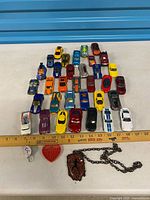Group of die-cast toy cars arranged in grid with ruler, necklace and keychains shown