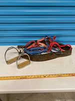 Bundle of stirrups, leather strap, blue and red reins on table with ruler for scale