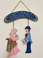 Full view of painted wooden welcome sign showing arch panel, two suspended figures and jute hanger