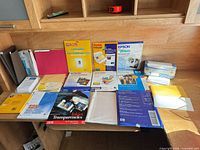 Overview of desk showing multiple unopened and opened packs of Epson, Kodak, HP, IBM and other specialty inkjet papers, transparencies, transfer sheets and envelopes